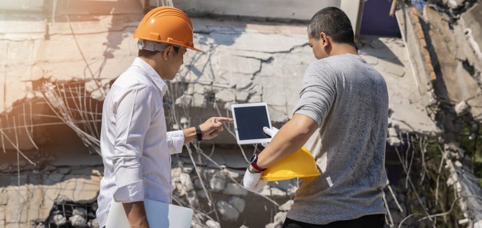 Two men in hardhats looking at a tablet in front of a building being demolished